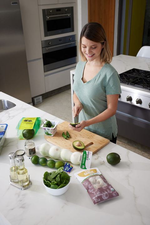 A shopper at home with EE Print Pack packaging on the counter: mesh-bagged limes and onions, a poly bag of peppers and a garlic pouch